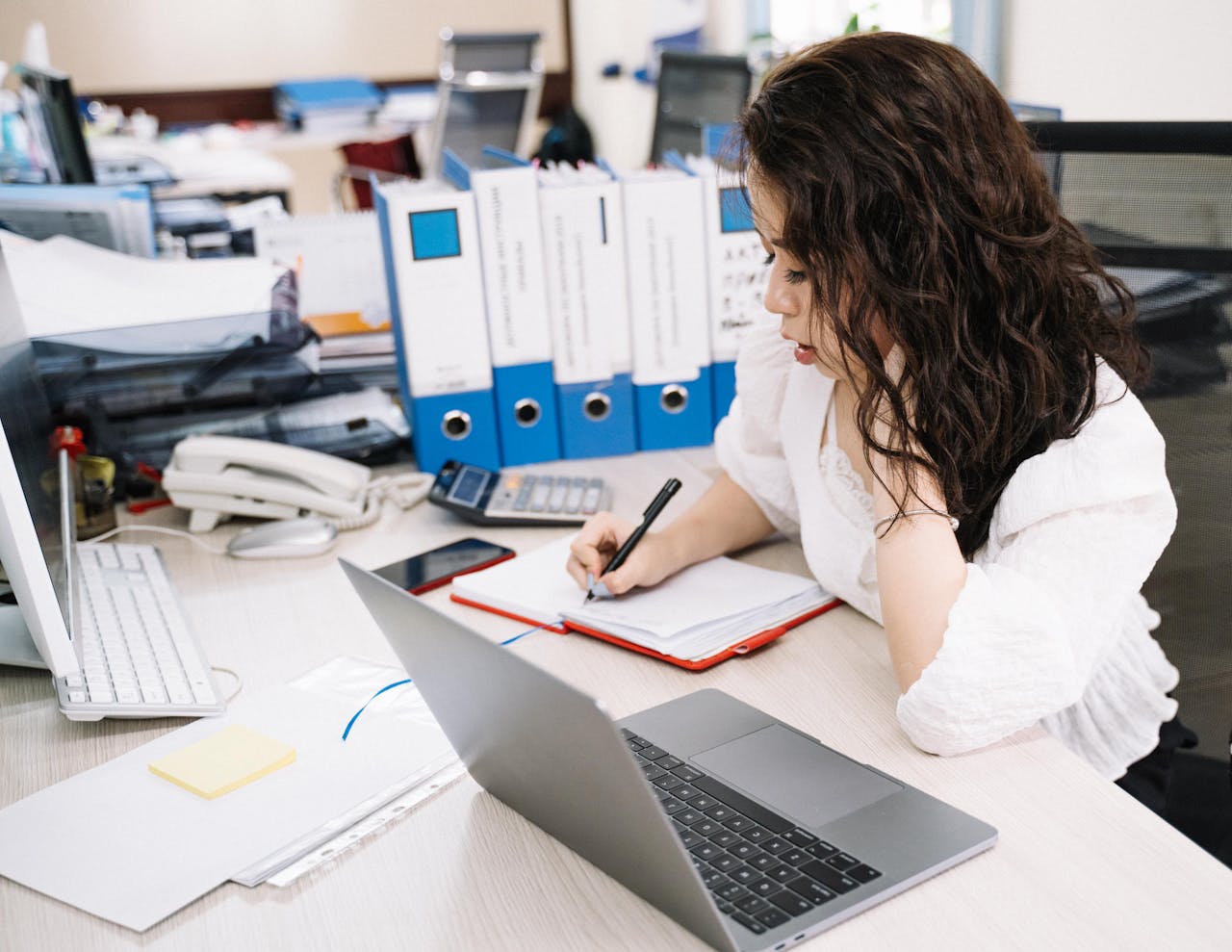 An office worker takes notes while working at a desk with a laptop and documents.